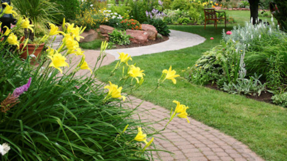 A landscaped garden with yellow flowers, curved stone paths, green grass, and a wooden bench surrounded by colorful plants.