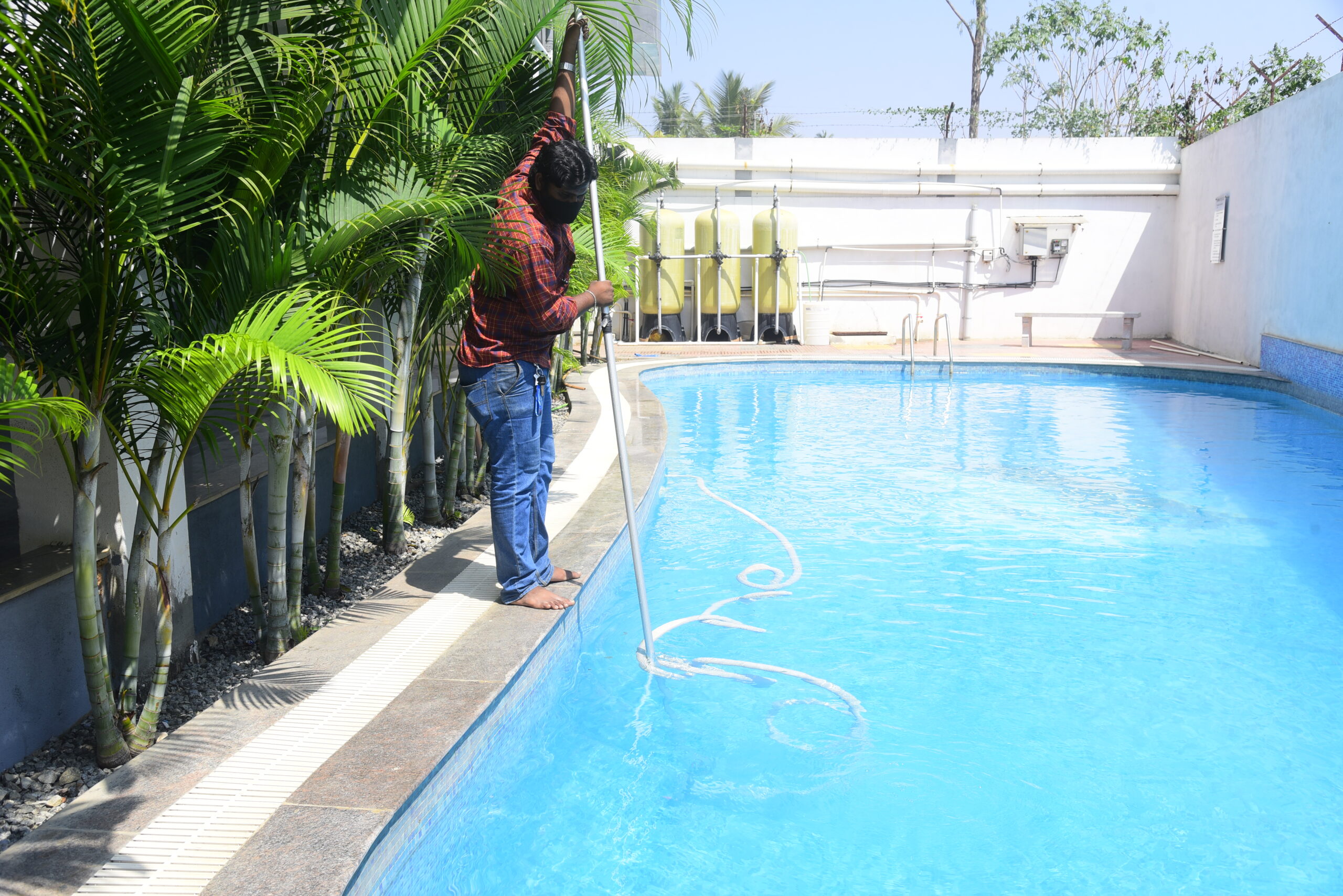 A worker cleans a swimming pool with equipment beside palm plants, representing Swimming pool Maintenance Services in Bangalore