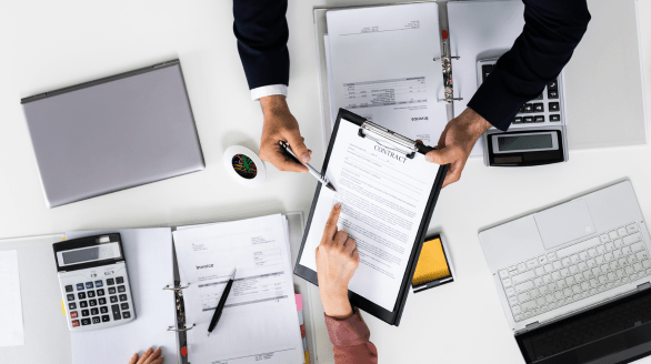 Two people reviewing and discussing a contract document on a clipboard surrounded by invoices, binders, calculators, and a laptop.