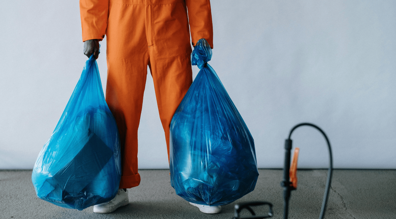 Soft services staff in orange uniform holding blue garbage bags and a cleaning sprayer nearby.